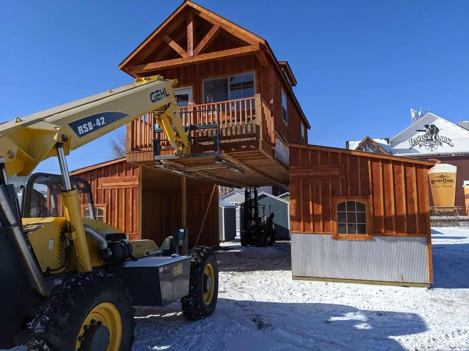 A yellow telehandler lifting a two-story wooden cabin with a balcony into place on a snowy lot