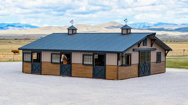 34×36 two-piece horse barn from the front: tan siding, black Dutch doors, two cupolas and a black metal roof, with mountains behind