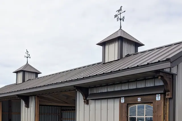 Two cupolas with horse weathervanes above a standing-seam metal roof, with bracketed eaves and a windowed door below