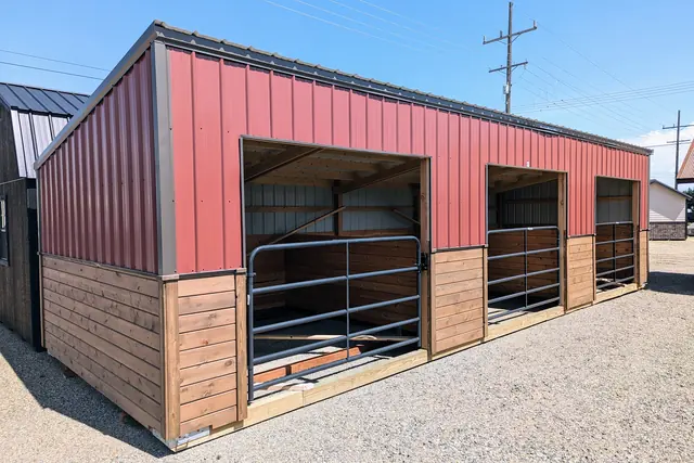Long red metal stable with three open stalls, each with a steel gate, above wood lower walls, on a gravel lot