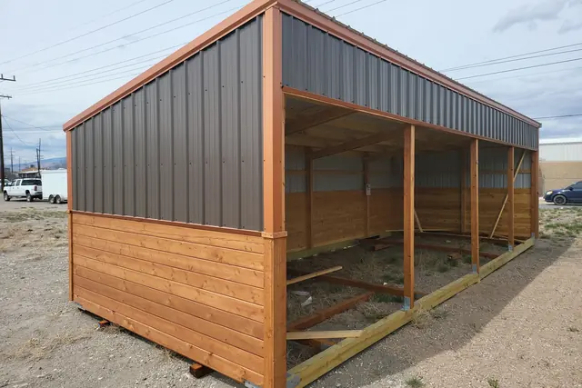 Long open-front run-in shelter with charcoal metal upper walls, copper-colored trim and wood lower walls, on a dirt lot