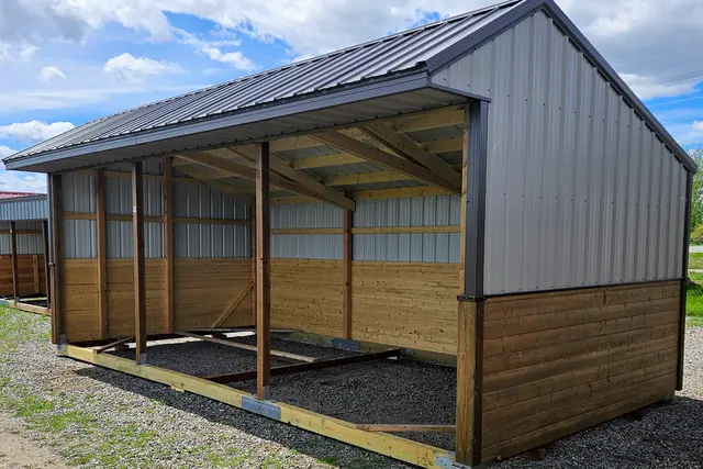 Open-front run-in shelter with a dark gabled metal roof and a gray gable end, on gravel with grass at its edge