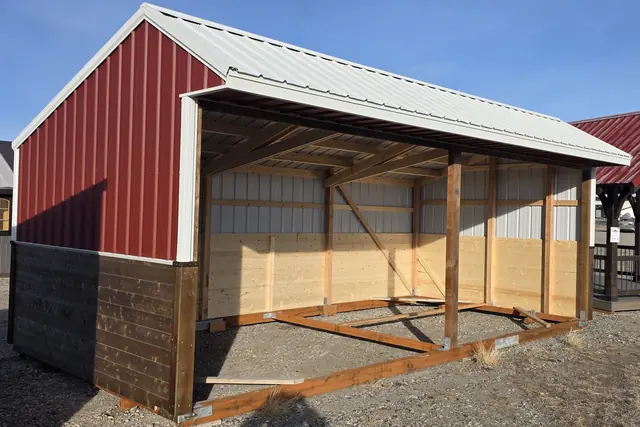 Open-front run-in shelter with a red metal gable end, a white metal roof and a wood-paneled back wall, on gravel