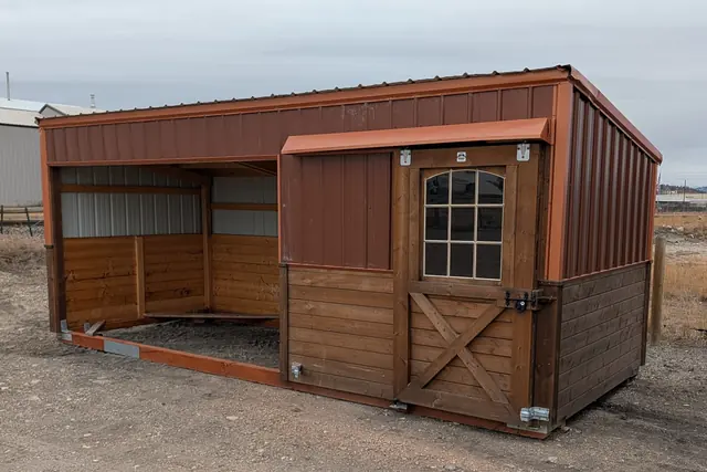 Open-front shelter with rust-colored metal upper walls and a windowed door with a cross-braced lower panel, on a dirt lot