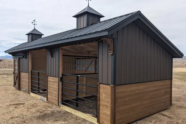 Stable with charcoal metal siding, two cupolas, a black metal roof and black steel gates on open stalls, in a dry field with hills behind