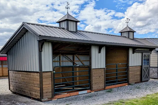 Gray stable with two cupolas, two stalls with green steel gates, wood lower walls and a gray cross-braced door, on a gravel lot