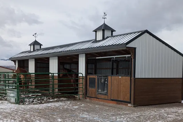 Stable with a gray roof and two cupolas, a stall with a black grille front, and two horses in green pipe pens beside it on snowy ground