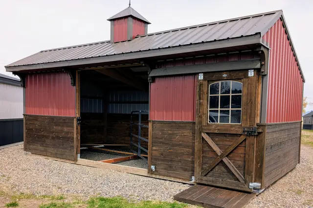 Red metal stable with a cupola and weathervane, an open stall and a windowed cross-braced door with a wooden ramp, on gravel and grass