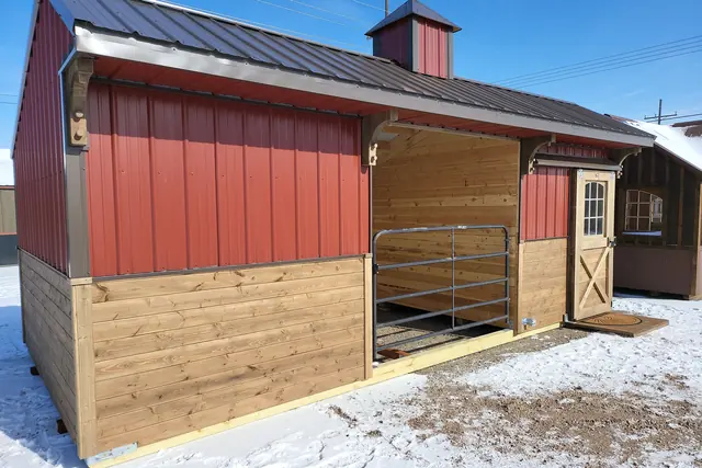 Red metal stable with a cupola, a steel gate across an open stall and a windowed door, standing in snow
