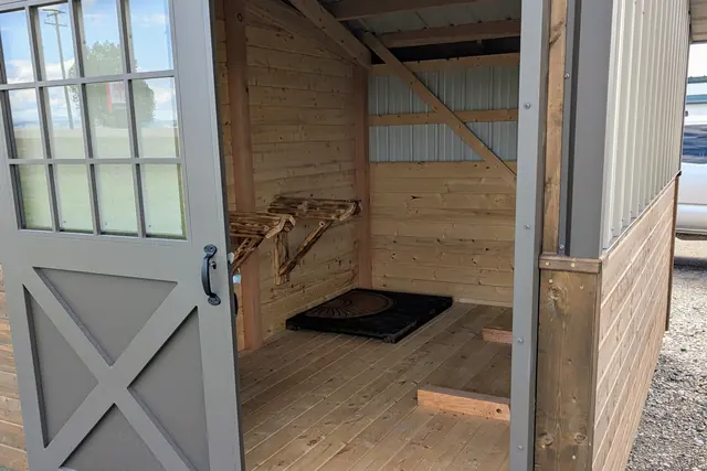 A tack room seen past an open gray cross-braced door with a many-paned window, with wooden saddle racks, a black mat and a plank floor