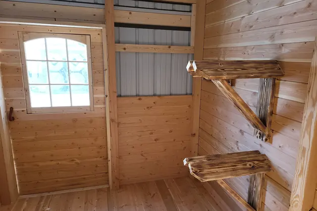Inside a small tack room: wooden saddle racks on a plank wall, an arched window and a plank floor