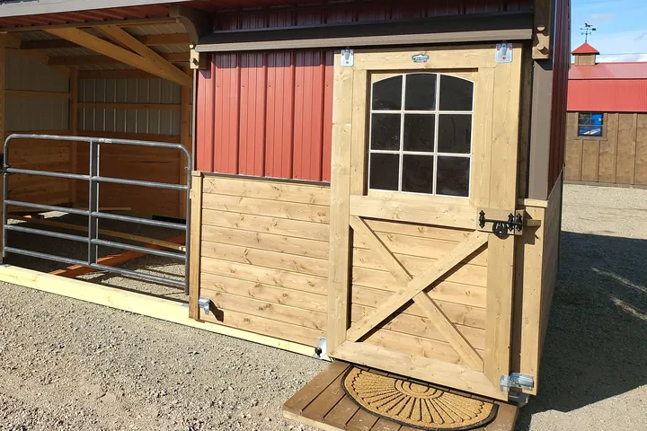 Close view of a windowed door with a cross-braced lower panel and black latch hardware, on a red metal stable
