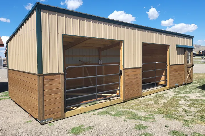 Long stable with tan metal upper walls, green trim and two open stalls with steel gates, on gravel and grass